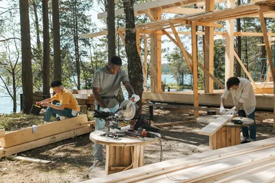 Team of diverse workers engaged in carpentry at a forested lakeside construction site.