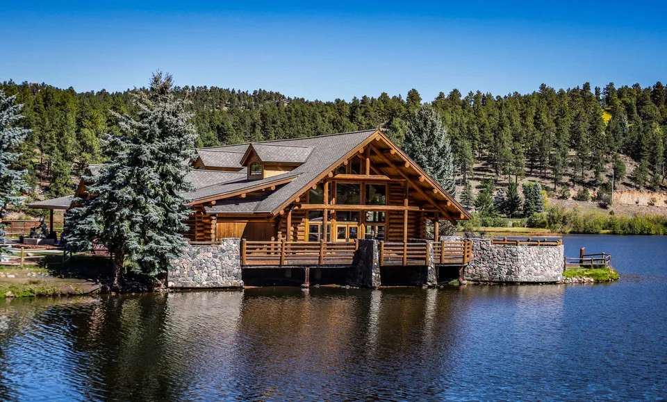 Beautiful wooden cabin by a serene lake surrounded by lush forest and clear blue sky.
